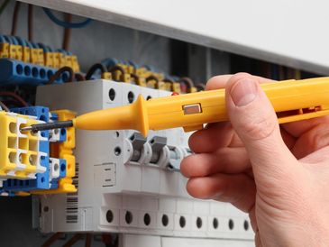 Hand holding a yellow voltage tester near electrical wiring and circuit breakers.