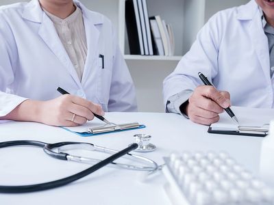 Two doctors in white coats writing on clipboards with medical tools in the foreground.