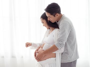 A couple lovingly embraces the woman's pregnant belly in a bright room.
