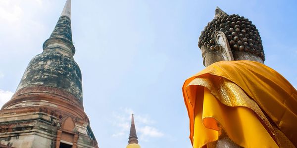 Buddha statue draped in orange robe near ancient pagodas under blue sky.