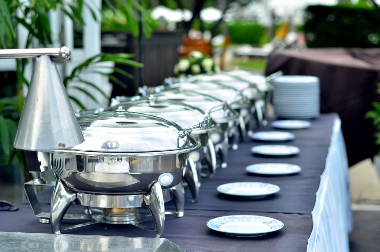 A row of shiny chafing dishes and plates on a buffet table.