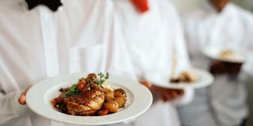 Waiters in white shirts serving plates with grilled chicken and vegetables.