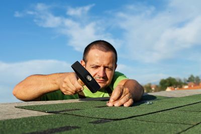 Man installing green roofing shingles with a hammer under a blue sky.