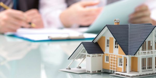 A miniature house model on a table with two people discussing documents in the background.