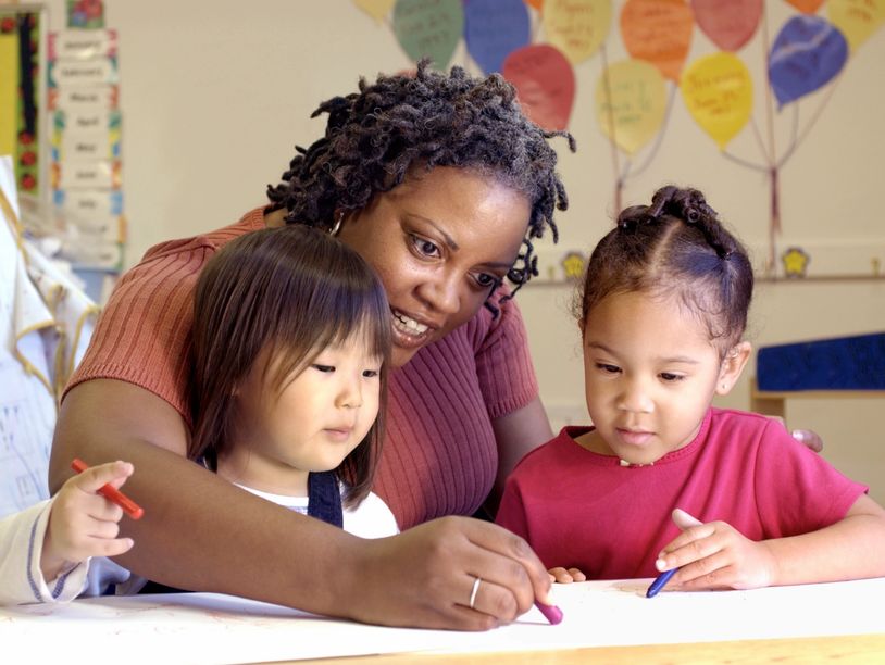 A teacher sitting between two toddlers helping them color on a paper