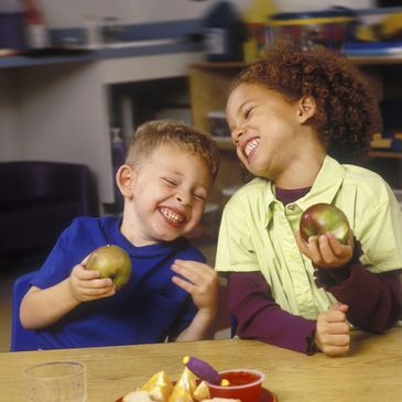 Two toddlers laughing eating apple sitting on chairs
