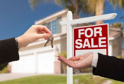 home seller passing keys to buyer just hands showing, next to for sale sign blurred house background