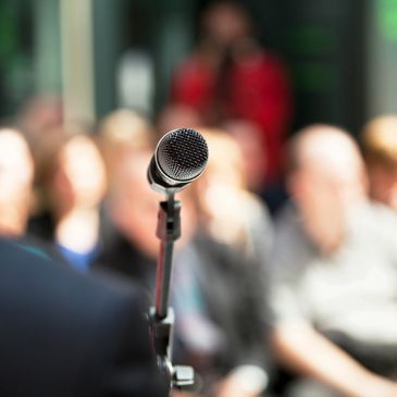 back shoulder of person standing at a microphone looking at a crowd of seated people