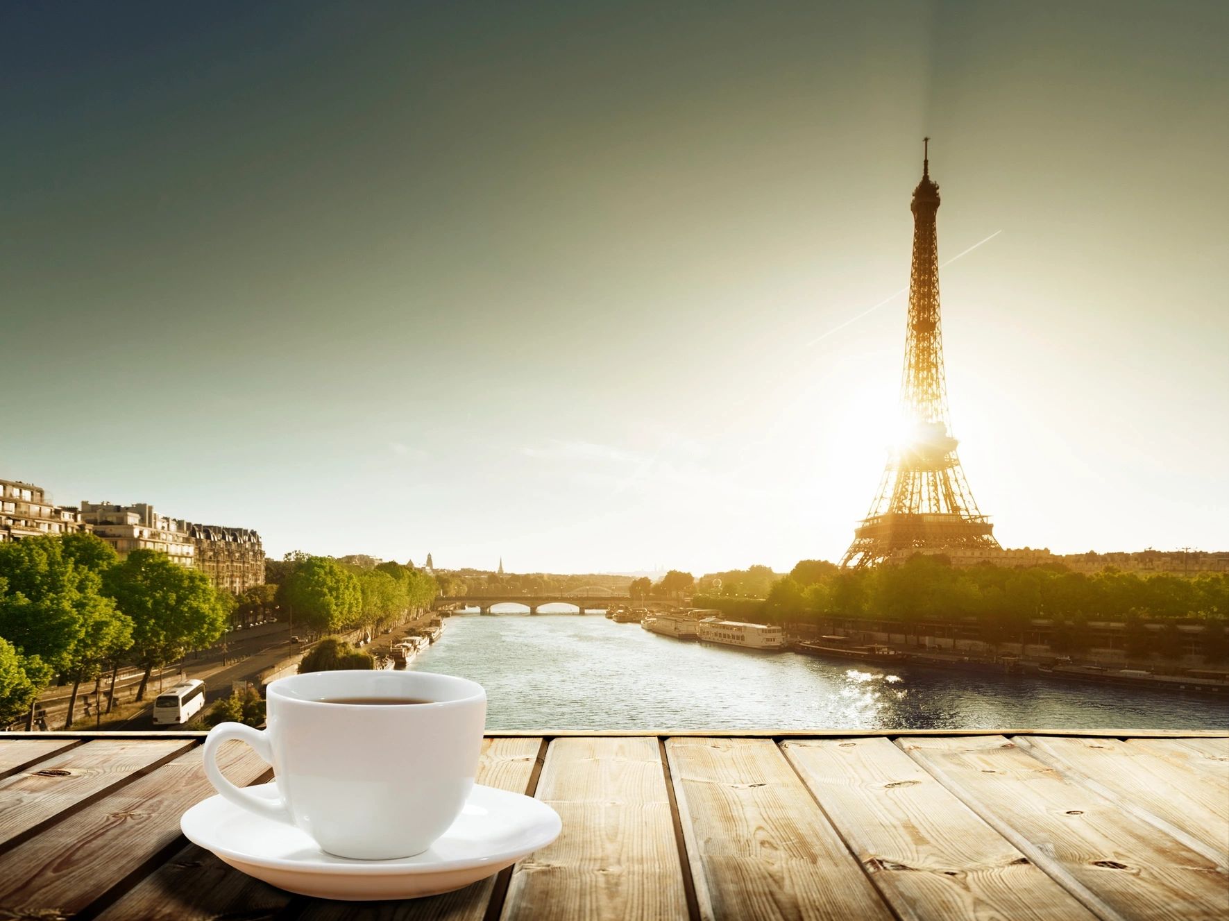 A cup of coffee on a wooden table with the Eiffel Tower and a river at sunrise.