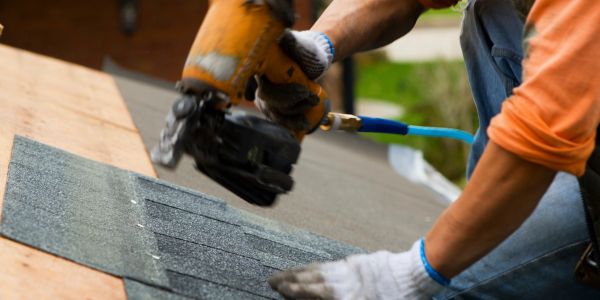 Worker installs shingles on a roof using a nail gun.