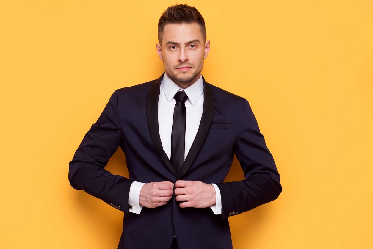 A gentleman in a black suit and tie and white shirt, buttons the top button of his blazer in front of a yellow wall.