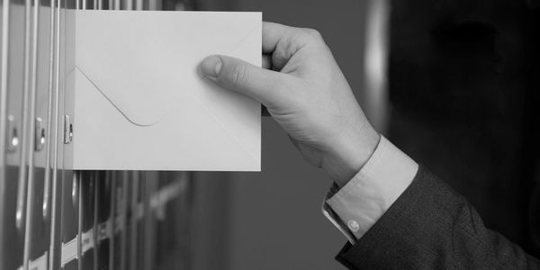 Hand inserting an envelope into a mailbox slot in black and white.
