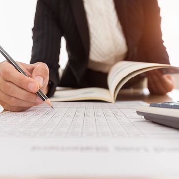 A woman in professional attire is sitting at a desk. The focus is on paperwork in the foreground.