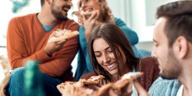 Friends laughing and eating pizza together indoors.