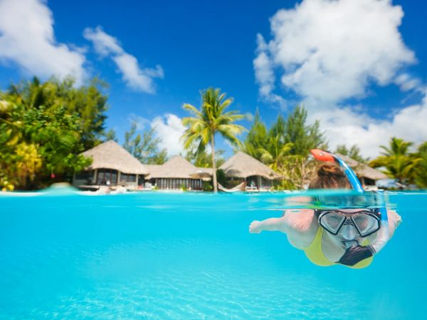 Person snorkeling in clear blue water near tropical huts.