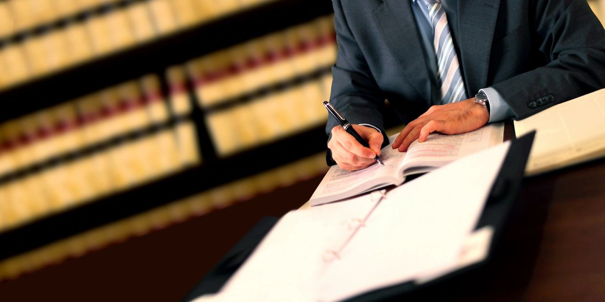 Person in suit writing in book at desk with law books in background.