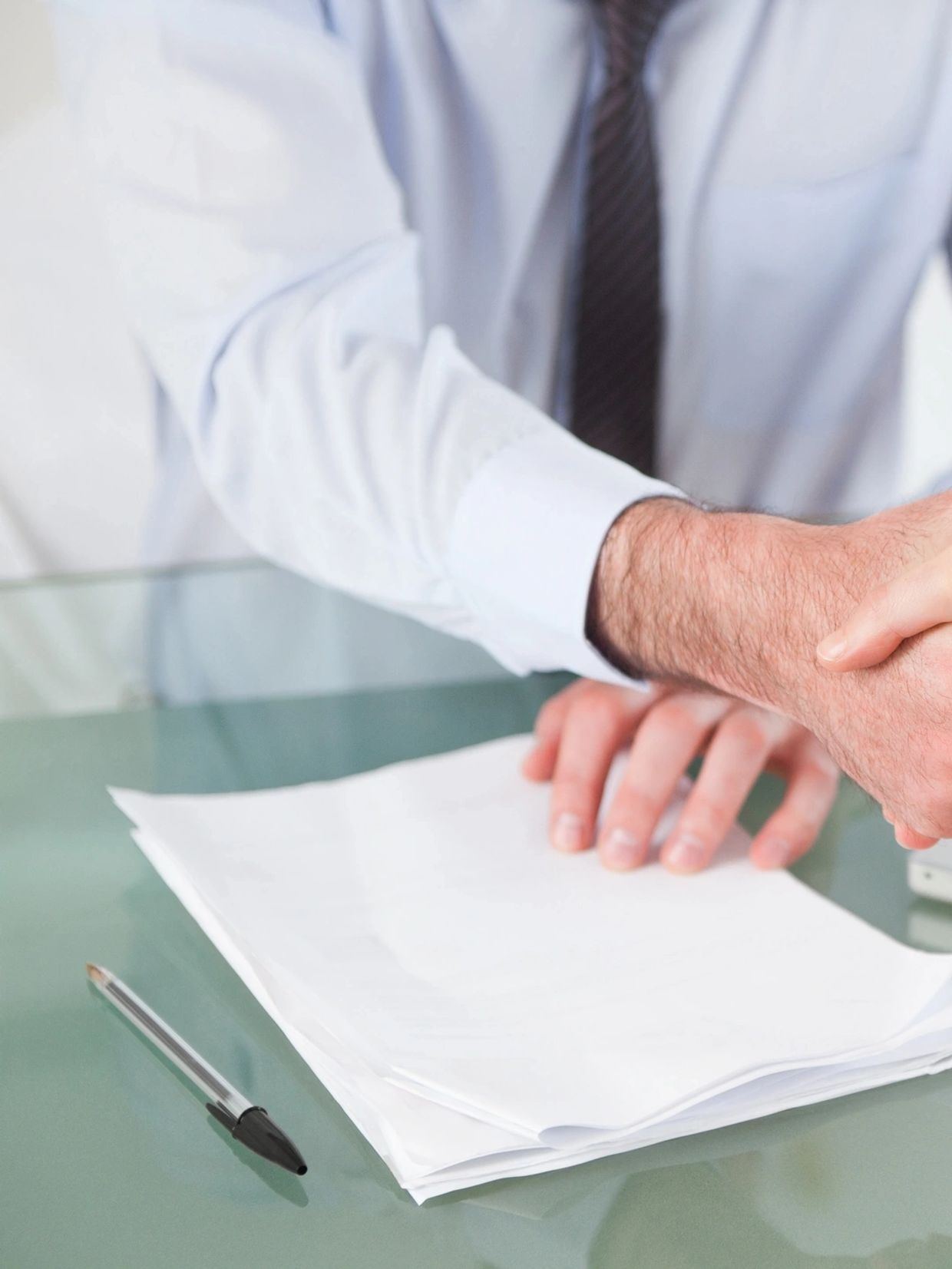 Two people shaking hands over signed documents on a glass table.