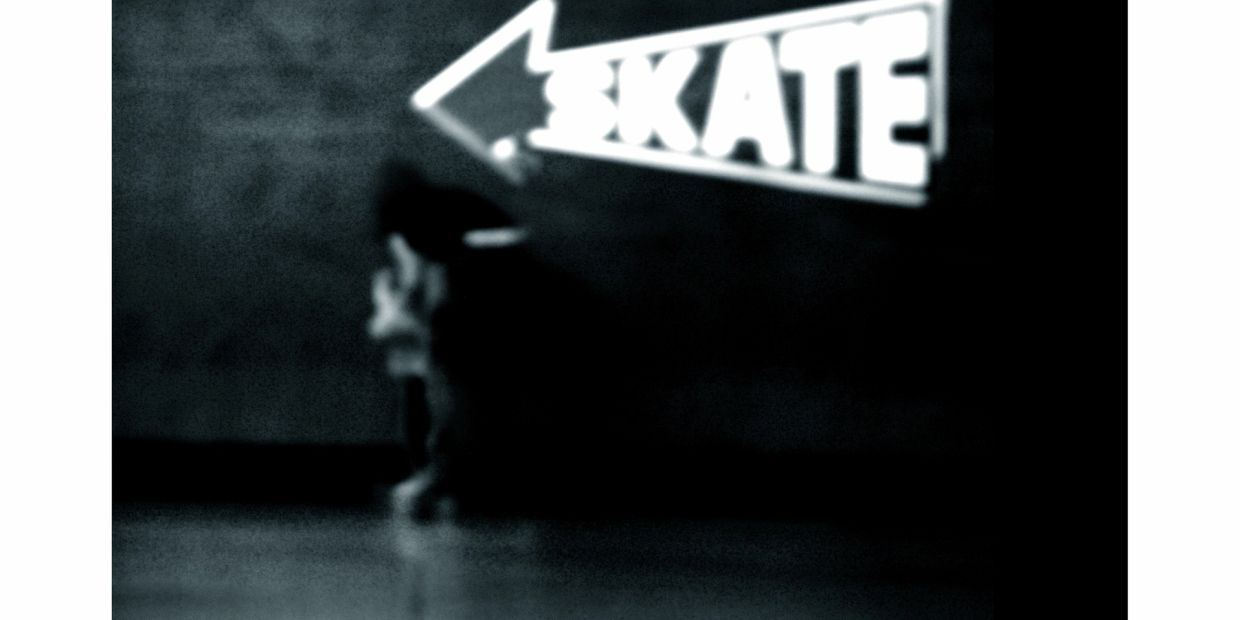 Blurry black-and-white image of a person sitting under a neon "SKATE" sign.