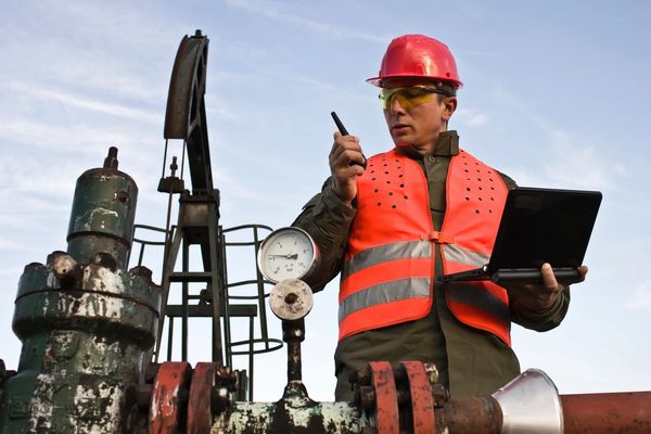Engineer in safety gear inspects industrial equipment using a laptop and walkie-talkie.