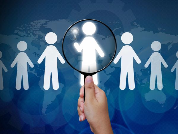 Magnifying glass highlighting a figure with a light bulb idea among others.