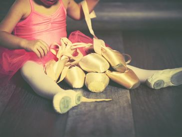 A young ballerina sitting on the floor surrounded by ballet pointe shoes.