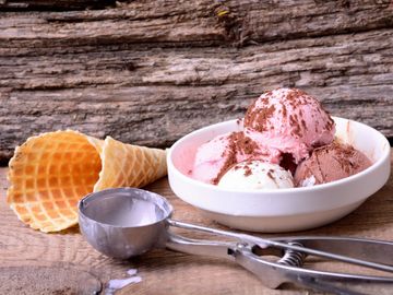 Bowl of assorted ice cream scoops with waffle cones and an ice cream scoop on a wooden surface.