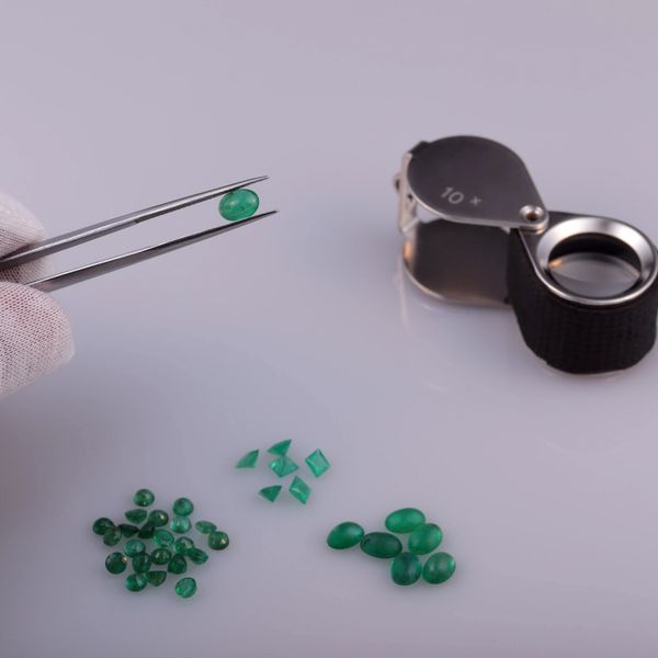 Green gemstones grouped by shape on the table with a magnifying glass and one held up by a tweezer. 