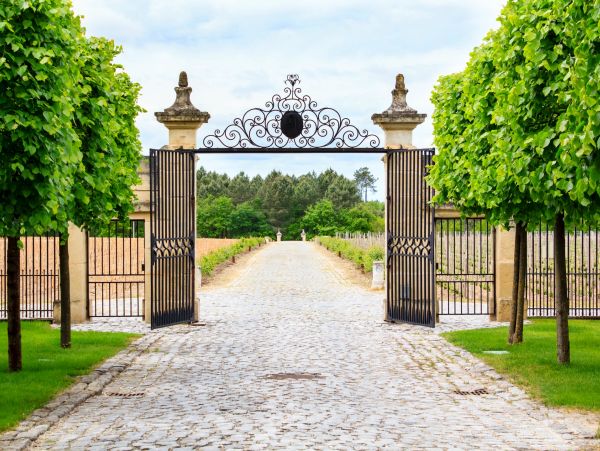 Open wrought iron gates lead to a tree-lined cobblestone path.