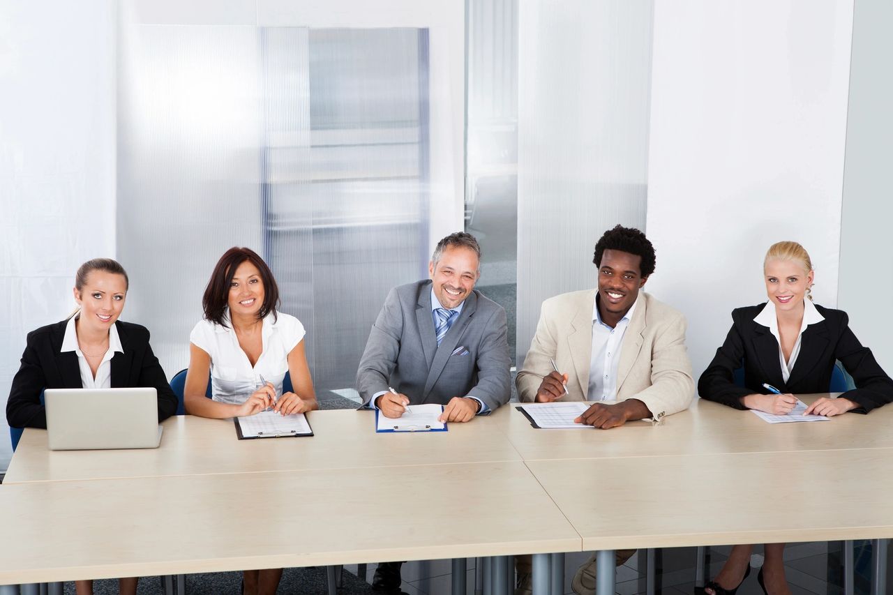 A panel of people smile as four take notes on their clipboards and one types on her laptop.