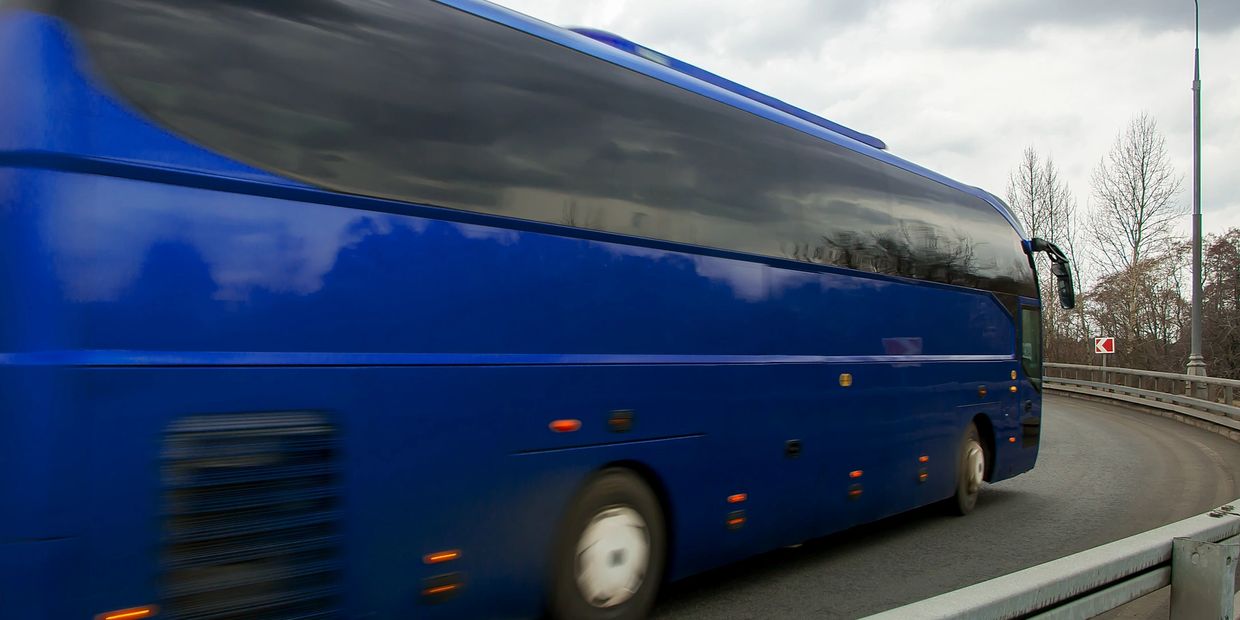 Blue coach bus driving on a curved road under cloudy sky.