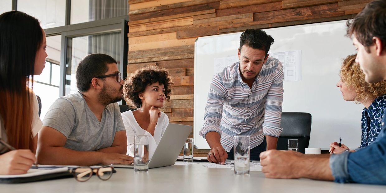 A team of people sitting around a table, looking up at the manager leaning over the table