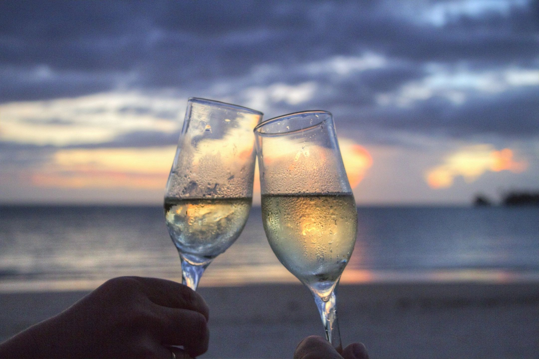 Two champagne glasses being clinked together while on a beach during sunset