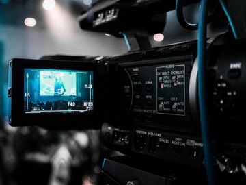 Close-up shot of a professional video camera capturing a live event, with the video camera's screen showing the event in progress. The background is slightly blurred, emphasizing the camera and its screen.