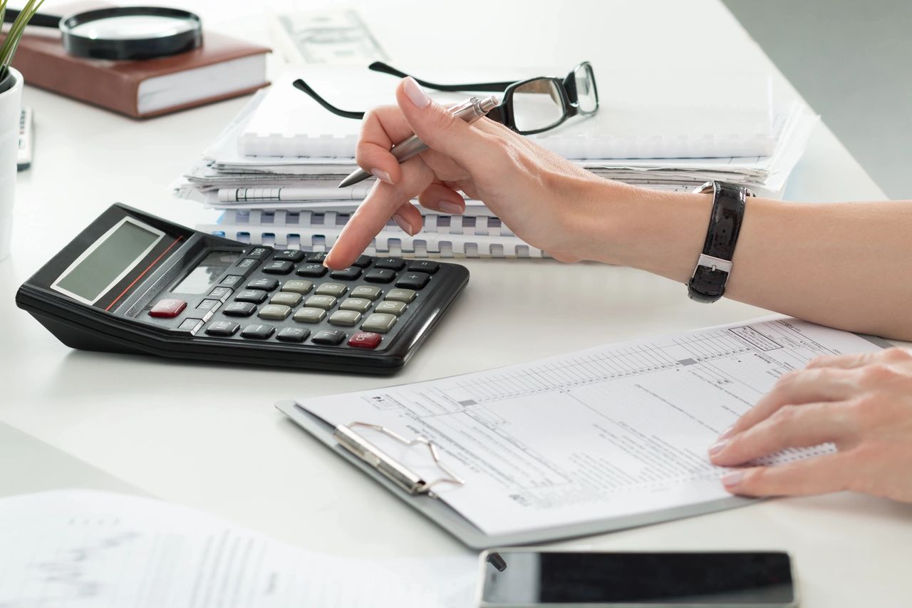 A person keys in figures into a calculator as they sit at their desk with a clipboard, eye glasses and cell phone nearby.