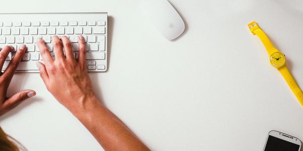 Hands typing on a white keyboard with a yellow watch and smartphone nearby.