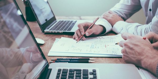 Two people sitting in front of laptops talking over pieces of paper with pens in hands.