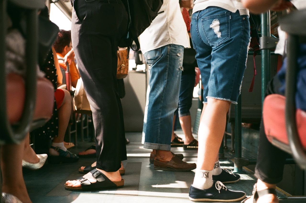 People standing in a bus, showcasing diverse footwear styles, including sandals, sneakers, and casual shoes, reflecting everyday commuting in a public transport setting.