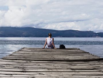 A person sitting on a wooden dock overlooking a calm lake with mountains in the background.