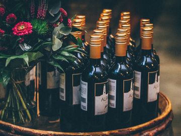 Rows of wine bottles with golden caps on a wooden surface beside flowers.