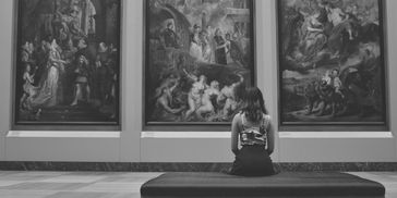 A woman sits alone in an art gallery, observing large classical paintings.