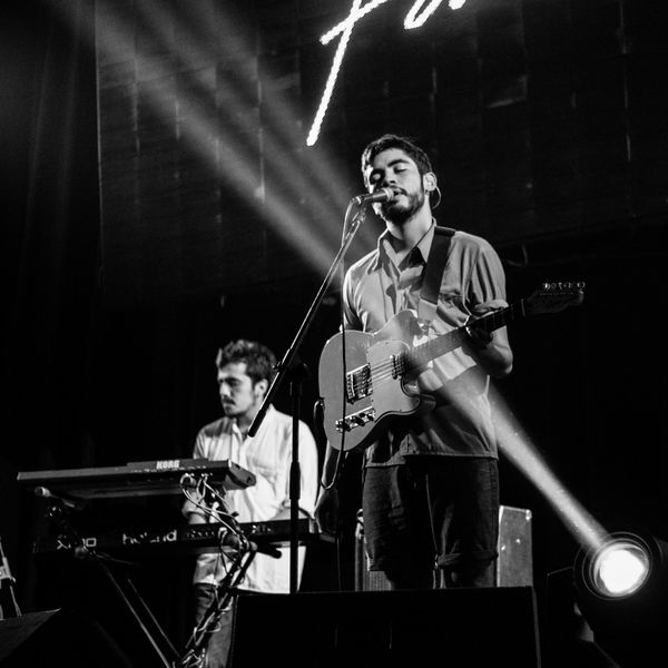 Black and white image of a musician singing and playing guitar with a keyboard player in the background.