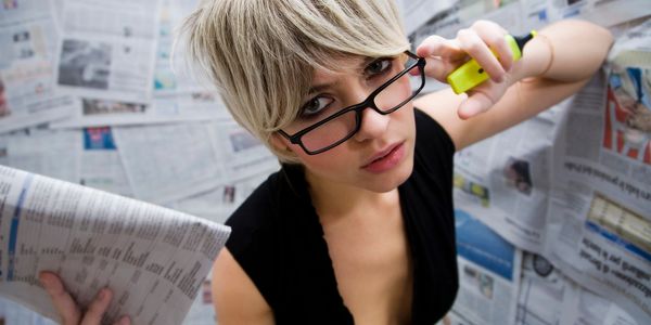 Woman with short blonde hair holding a newspaper and adjusting glasses.