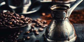 Close-up of a metal coffee pot surrounded by coffee beans on a dark surface.