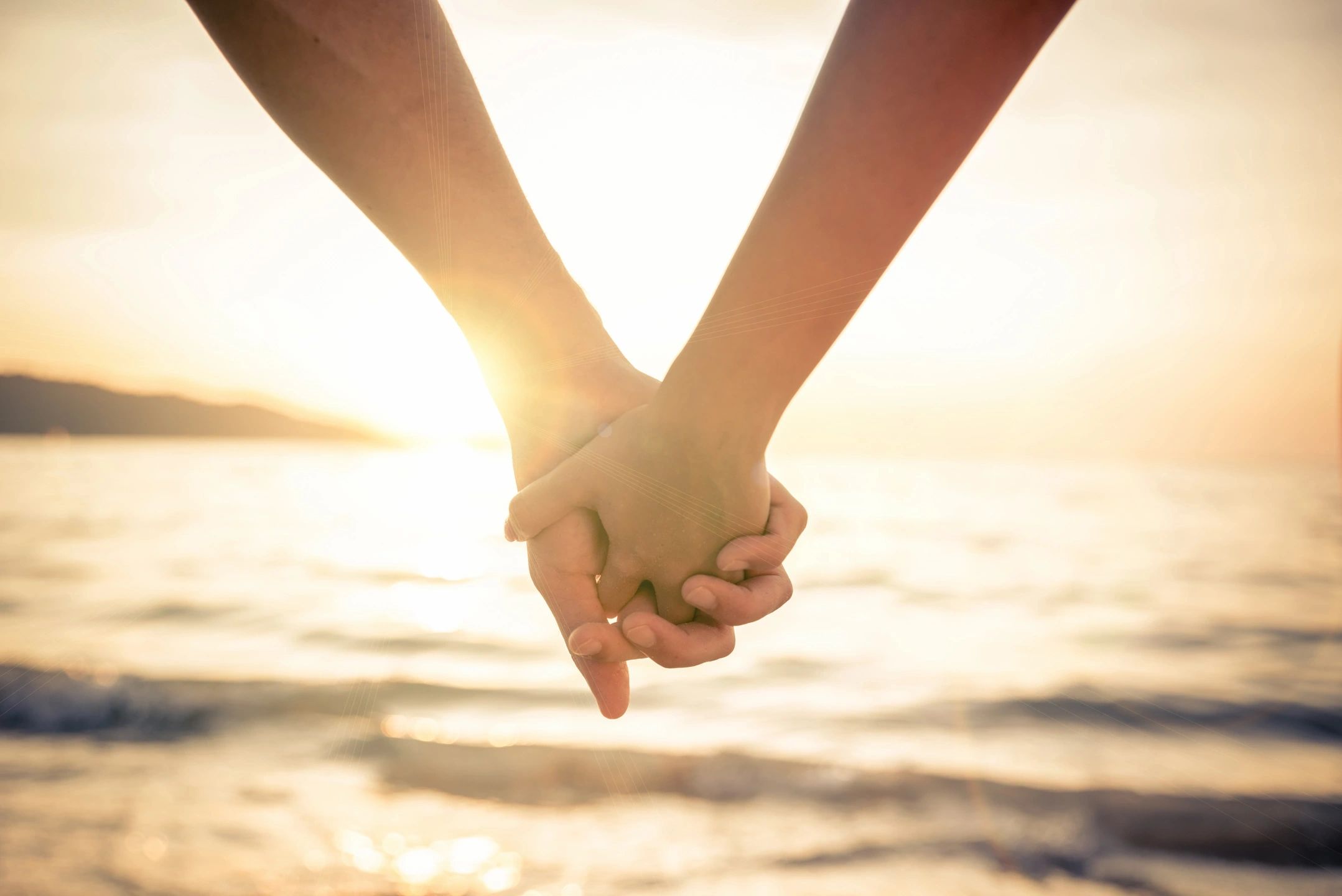 Two hands holding each other with the sun setting and the ocean in the background.