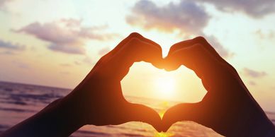 A photograph of two hands forming a heart shape with an ocean sunset in the background.