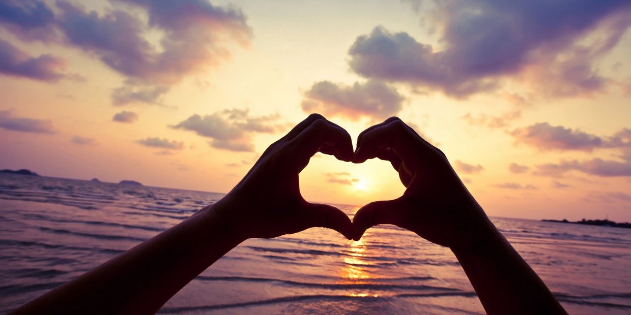 Two hands forming a heart shape raised towards the sunset sky and ocean.