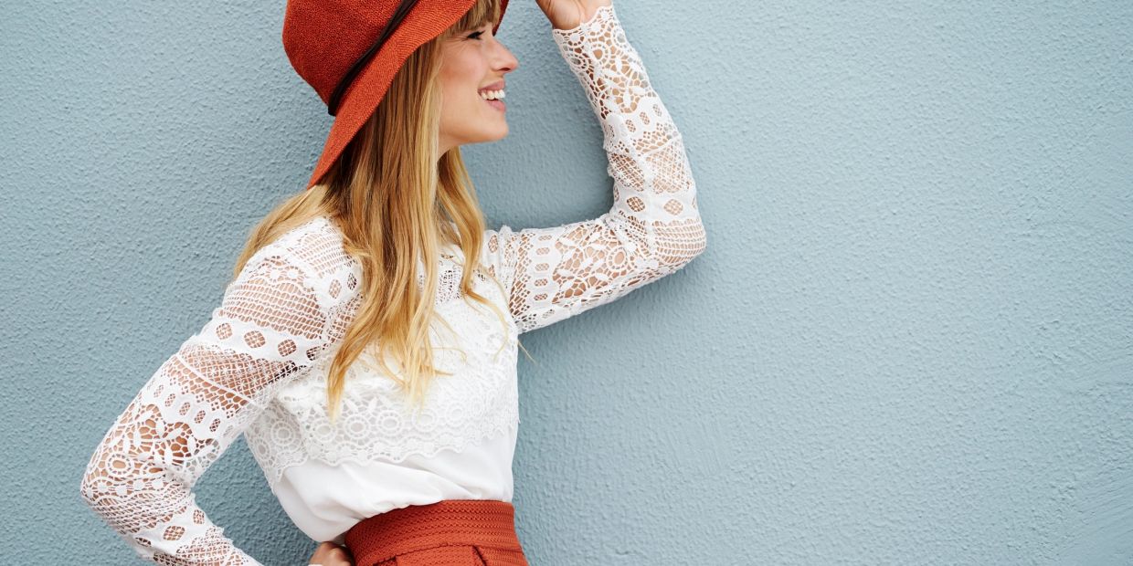 Woman in lace top and orange hat smiling against blue wall.