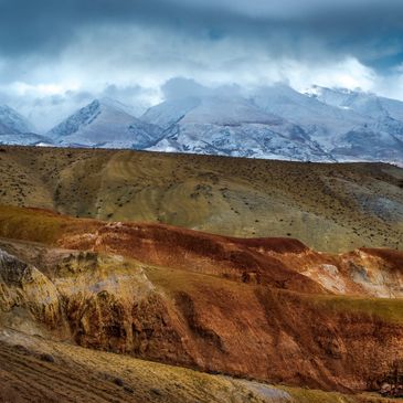 Colorful layered hills with snow-covered mountains and clouds in the background.