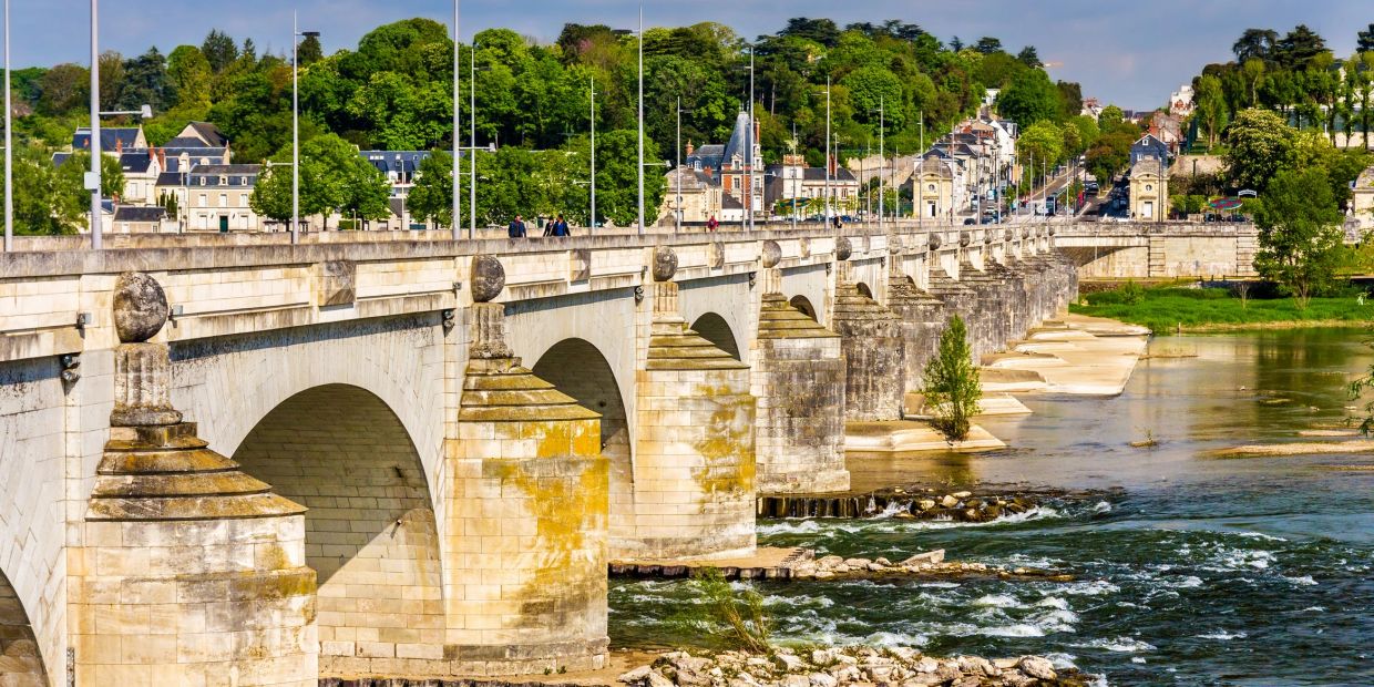 Historic stone bridge over a flowing river with green trees and buildings in the background.