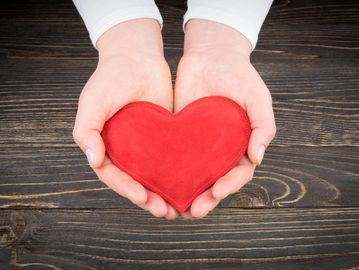 Hands holding a red heart on a wooden surface.